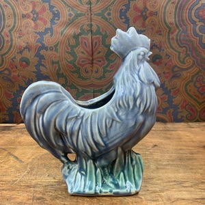 May include: A ceramic rooster planter in shades of blue and green. The rooster is standing on a base of green grass. The planter has a hollowed-out back for holding plants or flowers. The background is a patterned fabric.