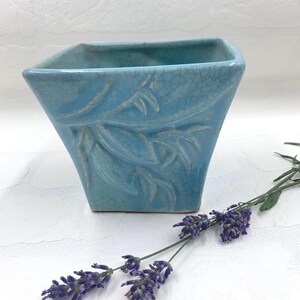 May include: A square-shaped turquoise ceramic planter featuring a crackle glaze finish and a raised relief pattern of leaves on the front side, accompanied by a sprig of purple lavender flowers placed next to the base of the container.