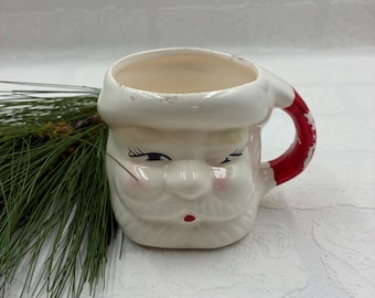 Könnte beinhalten: Ein Keramikbecher in Form eines Weihnachtsmann-Gesichts mit weißem Bart, Schnurrbart und Hutkrempe. Der Becher hat einen roten Henkel, aufgemalte blaue Augen mit langen Wimpern, rosa Wangen und einen kleinen roten Mund. Farbabrieb am Henkel.
