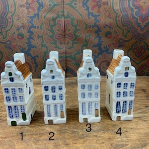 May include: Four ceramic miniature house figurines. Each house is white with blue window and door details, and a brown roof. The houses are numbered 1 through 4. The background is a patterned fabric.