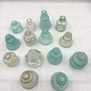 May include: A collection of thirteen small glass bottles in various shapes and sizes, featuring clear and light blue-tinted glass. Each bottle has a narrow neck and a circular opening at the top, with some displaying rounded, conical, or hexagonal bases.
