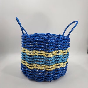 Könnte beinhalten: Ein blauer und gelber Korb aus Seil mit zwei Griffen. Der Korb ist aus blauem und gelbem Seil geflochten und hat eine natürliche braune Seilauskleidung.