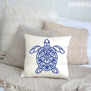 K&ouml;nnte beinhalten: Ein wei&szlig;es Kissen mit einem blauen Mandala-artigen Meeresschildkr&ouml;ten-Design. Das Kissen liegt auf einem Bett mit einer wei&szlig;en Decke und einem braunen &Uuml;berwurf.