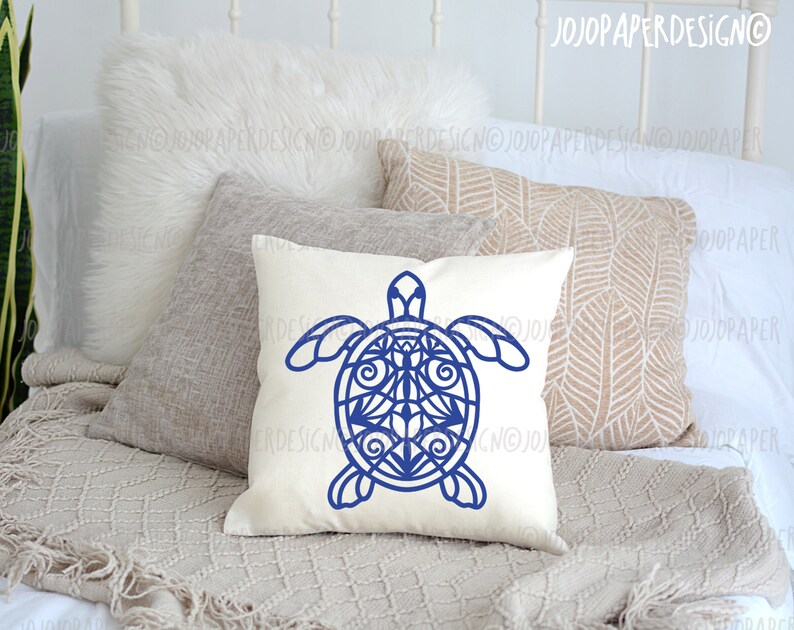 K&ouml;nnte beinhalten: Ein wei&szlig;es Kissen mit einem blauen Mandala-artigen Meeresschildkr&ouml;ten-Design. Das Kissen liegt auf einem Bett mit einer wei&szlig;en Decke und einem braunen &Uuml;berwurf.