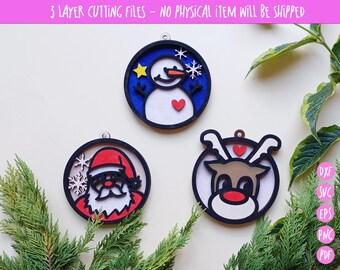 May include: Three layered Christmas ornament designs for cutting. The designs include a snowman with a red heart, a reindeer with a red nose, and Santa Claus with a white beard. The ornaments are on a white background with green foliage.