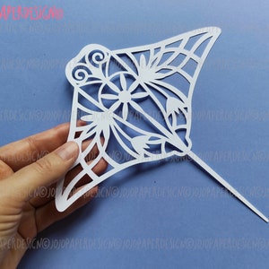 K&ouml;nnte beinhalten: Wei&szlig;er Papierschnitt einer Manta Rochen mit filigranen Blumenmustern. Der Ausschnitt wird in einer Hand gehalten und hat einen d&uuml;nnen Stock am Boden.