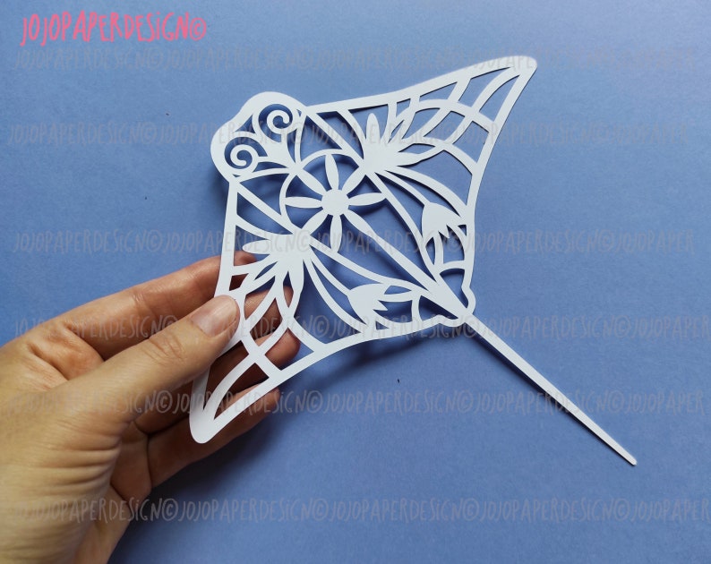 K&ouml;nnte beinhalten: Wei&szlig;er Papierschnitt einer Manta Rochen mit filigranen Blumenmustern. Der Ausschnitt wird in einer Hand gehalten und hat einen d&uuml;nnen Stock am Boden.