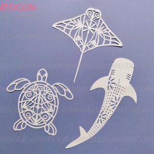 K&ouml;nnte beinhalten: Drei wei&szlig;e Papierschnitte von Meerestieren: ein Mantarochen, eine Schildkr&ouml;te und ein Walhai. Die Ausschnitte sind filigran und detailliert, mit einem zarten Blumenmuster.