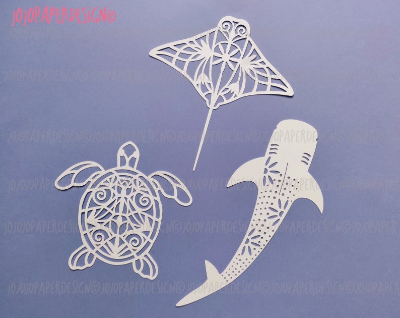 K&ouml;nnte beinhalten: Drei wei&szlig;e Papierschnitte von Meerestieren: ein Mantarochen, eine Schildkr&ouml;te und ein Walhai. Die Ausschnitte sind filigran und detailliert, mit einem zarten Blumenmuster.