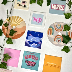 K&ouml;nnte beinhalten: Eine Sammlung quadratischer und runder dekorativer Magnete mit verschiedenen Designs und Texten. Die Magnete zeigen S&auml;tze wie "CALL ME IF YOU GET LOST" und "HEAVYWEIGHT GIRLFRIEND", zusammen mit einem "MARVEL"-Logo und Albumcovern.