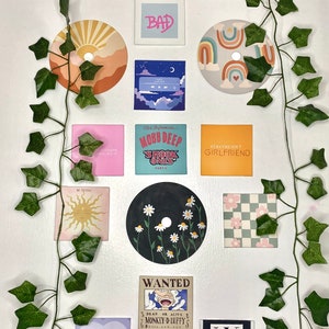 K&ouml;nnte beinhalten: Eine Sammlung dekorativer Wandkunst, darunter bemalte CDs, quadratische Drucke und ein Marvel-Logo. Die Kunstwerke zeigen verschiedene Designs, wie einen Sonnenuntergang, Regenbogen, G&auml;nsebl&uuml;mchen und Text. Gr&uuml;ne Efeuranken sind um die Kunstwerke drapiert.