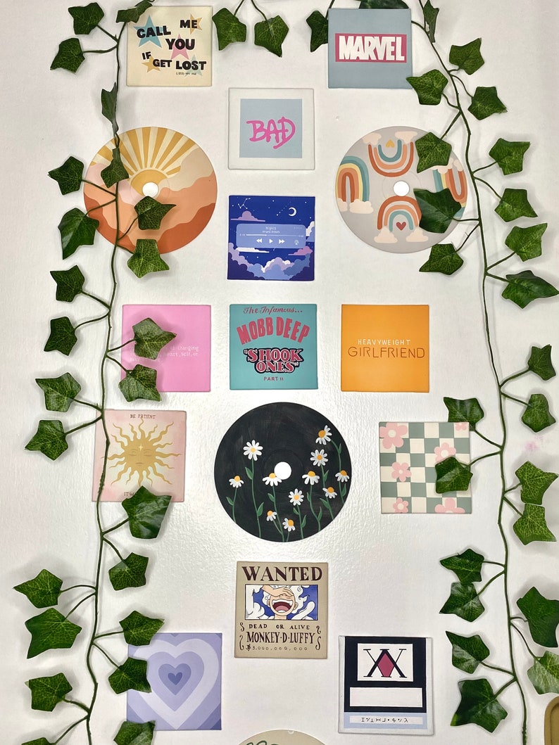 K&ouml;nnte beinhalten: Eine Sammlung dekorativer Wandkunst, darunter bemalte CDs, quadratische Drucke und ein Marvel-Logo. Die Kunstwerke zeigen verschiedene Designs, wie einen Sonnenuntergang, Regenbogen, G&auml;nsebl&uuml;mchen und Text. Gr&uuml;ne Efeuranken sind um die Kunstwerke drapiert.