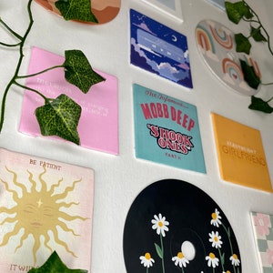 K&ouml;nnte beinhalten: Eine Sammlung bemalter Leinw&auml;nde und eine schwarze CD mit G&auml;nsebl&uuml;mchen verziert. Die Leinw&auml;nde zeigen verschiedene Designs, darunter Texte mit der Aufschrift "BAD", "MARVEL", "BE PATIENT" und "HEAVYWEIGHT GIRLFRIEND". Andere Leinw&auml;nde haben abstrakte Designs.