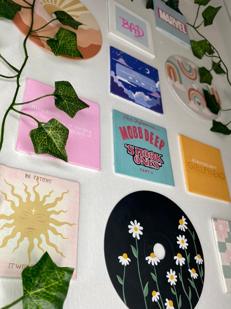 K&ouml;nnte beinhalten: Eine Sammlung bemalter Leinw&auml;nde und eine schwarze CD mit G&auml;nsebl&uuml;mchen verziert. Die Leinw&auml;nde zeigen verschiedene Designs, darunter Texte mit der Aufschrift "BAD", "MARVEL", "BE PATIENT" und "HEAVYWEIGHT GIRLFRIEND". Andere Leinw&auml;nde haben abstrakte Designs.