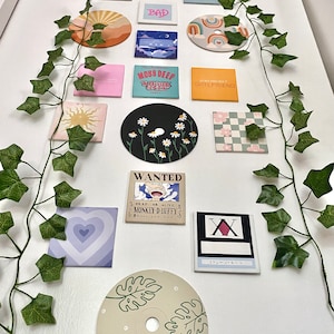 K&ouml;nnte beinhalten: Eine Sammlung dekorativer Magnete mit verschiedenen Designs und Texten, darunter eine Sonne, ein Herz und die Worte "CALL ME YOU GET LOST". Die Magnete sind auf einer wei&szlig;en Oberfl&auml;che angeordnet und mit k&uuml;nstlichem Efeu verziert.