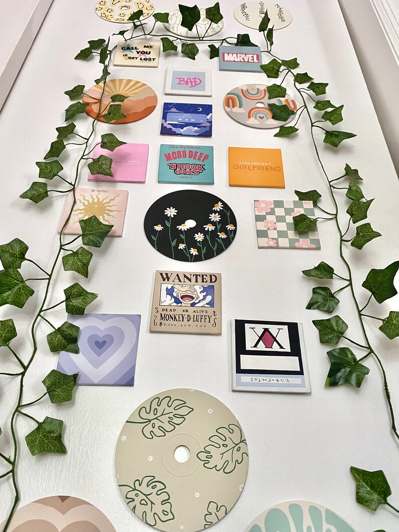 K&ouml;nnte beinhalten: Eine Sammlung dekorativer Magnete mit verschiedenen Designs und Texten, darunter eine Sonne, ein Herz und die Worte "CALL ME YOU GET LOST". Die Magnete sind auf einer wei&szlig;en Oberfl&auml;che angeordnet und mit k&uuml;nstlichem Efeu verziert.