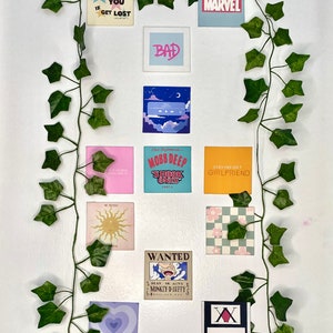 K&ouml;nnte beinhalten: Eine Anordnung quadratischer Kunstdrucke mit verschiedenen Designs und Texten, darunter "CALL ME IF YOU GET LOST," "BAD" und "MARVEL." Die Drucke sind an einer wei&szlig;en Wand mit k&uuml;nstlichem Efeu angebracht.