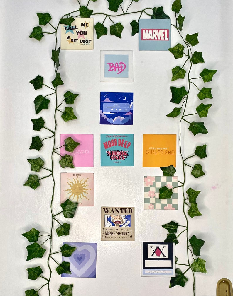 K&ouml;nnte beinhalten: Eine Anordnung quadratischer Kunstdrucke mit verschiedenen Designs und Texten, darunter "CALL ME IF YOU GET LOST," "BAD" und "MARVEL." Die Drucke sind an einer wei&szlig;en Wand mit k&uuml;nstlichem Efeu angebracht.