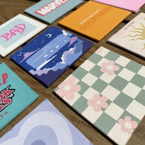 K&ouml;nnte beinhalten: Eine Sammlung quadratischer, handgemalter Leinw&auml;nde mit verschiedenen Designs. Die Designs umfassen Text, ein Schachbrettmuster mit rosa Blumen, eine Musikplayer-Oberfl&auml;che und das Marvel-Logo. Die Leinw&auml;nde sind auf einer Holzoberfl&auml;che angeordnet.