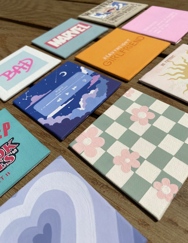 K&ouml;nnte beinhalten: Eine Sammlung quadratischer, handgemalter Leinw&auml;nde mit verschiedenen Designs. Die Designs umfassen Text, ein Schachbrettmuster mit rosa Blumen, eine Musikplayer-Oberfl&auml;che und das Marvel-Logo. Die Leinw&auml;nde sind auf einer Holzoberfl&auml;che angeordnet.