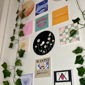 K&ouml;nnte beinhalten: Eine Sammlung dekorativer Wandkunst, darunter eine schwarze Schallplatte mit wei&szlig;en G&auml;nsebl&uuml;mchen, ein Quadrat mit dem Text "HEAVYWEIGHT GIRLFRIEND" und ein "WANTED"-Poster. Gr&uuml;ne Efeuranken sind um die Kunst drapiert.