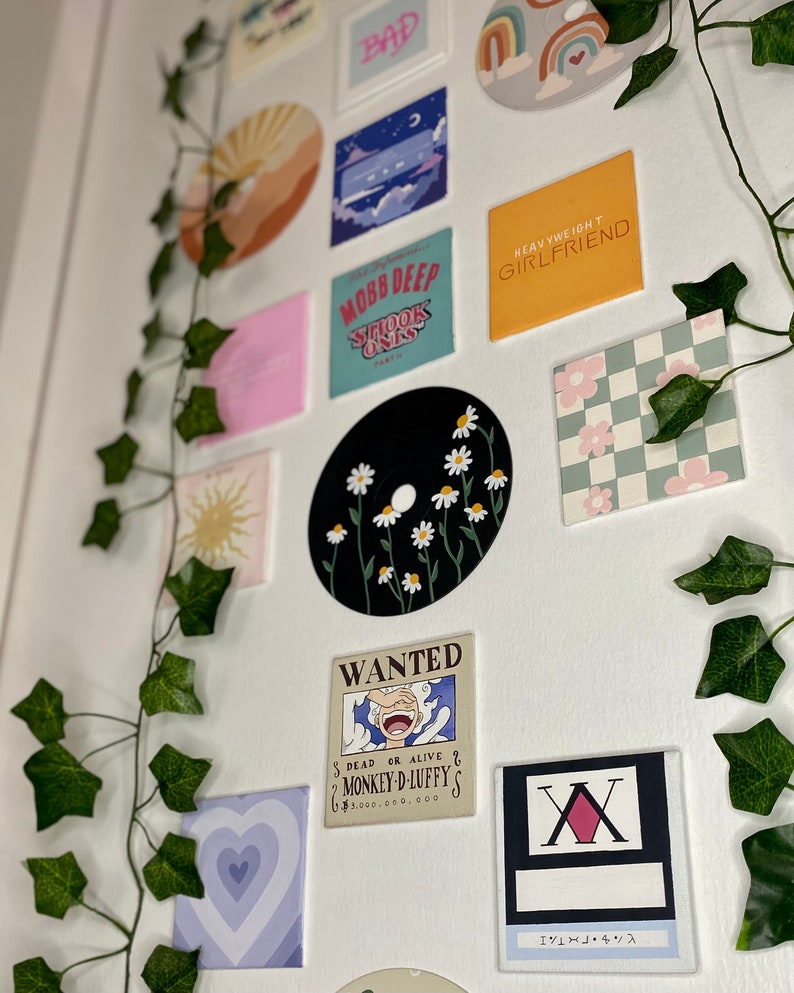 K&ouml;nnte beinhalten: Eine Sammlung dekorativer Wandkunst, darunter eine schwarze Schallplatte mit wei&szlig;en G&auml;nsebl&uuml;mchen, ein Quadrat mit dem Text "HEAVYWEIGHT GIRLFRIEND" und ein "WANTED"-Poster. Gr&uuml;ne Efeuranken sind um die Kunst drapiert.