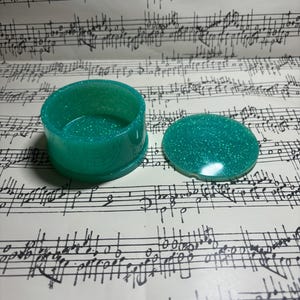 May include: A teal green glitter resin trinket box with a lid. The box is round and has a smooth, shiny finish.