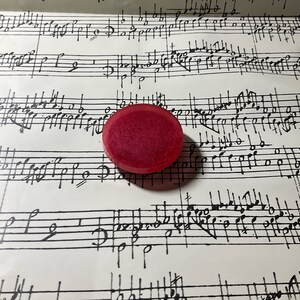 May include: A red, round, flat, decorative object with a slightly textured surface. The object is sitting on a sheet of music.
