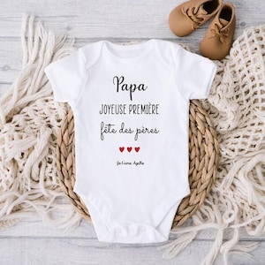 Peut inclure: Un body blanc pour bébé avec du texte noir qui dit "Papa Joyeuse Premiere Fete des Peres" avec trois cœurs rouges et le texte "Je t'aime Agathe".