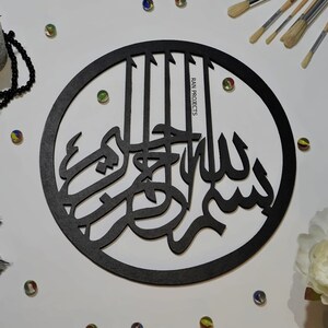 May include: Black wooden round wall decor with Arabic calligraphy that reads "Bismillah" in a circular frame. The text is in a decorative font and is centered in the frame. The frame is surrounded by a white background.