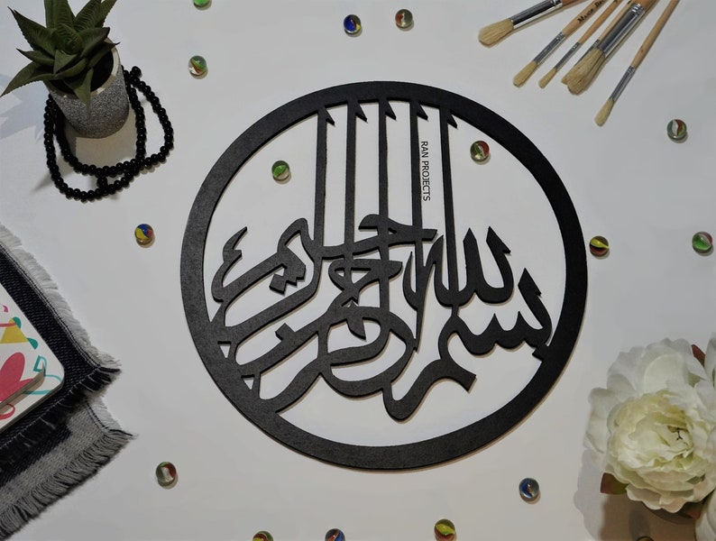 May include: Black wooden round wall decor with Arabic calligraphy that reads "Bismillah" in a circular frame. The text is in a decorative font and is centered in the frame. The frame is surrounded by a white background.