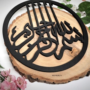 May include: A black wooden round sign with a circular cutout featuring the Arabic calligraphy for "Allah" in a bold, stylized font. The sign is mounted on a natural wood slice with the text "RAN PROJECTS" inscribed at the bottom.