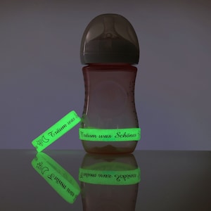 Könnte beinhalten: Eine rosa Babyflasche mit einem durchsichtigen Sauger und einem leuchtend grünen Band mit der Aufschrift "Träum was Schönes". Ein passendes leuchtend grünes Band mit dem gleichen Text befindet sich neben der Flasche. Der Text ist in schwarzer Schrift.