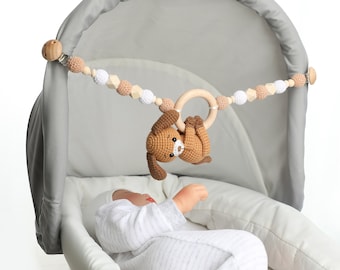 Pode incluir: Um brinquedo de cachorro de crochê marrom pendurado em um móbile de contas de madeira e um anel, que está preso a um berço de bebê cinza. O móbile é feito de contas de madeira, contas de crochê e um anel de madeira. Um bebê está deitado no berço.