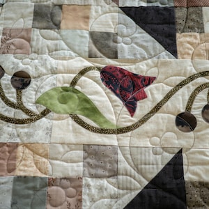 May include: A close-up of a quilt with a floral design. The quilt is made of various fabrics in shades of brown, beige, and green. A red flower with a green leaf is in the center of the quilt.