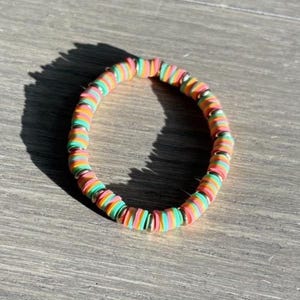 Puede incluir: Un brazalete colorido hecho de cuentas redondas apiladas. Las cuentas están superpuestas en tonos de rosa, naranja y verde, con un acento dorado.