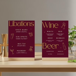 Może przedstawiać: Dwa bordowe znaki na stół ze złotym tekstem i ilustracjami. Jeden znak mówi "Libations" i wymienia koktajle. Drugi mówi "Wine" i "Beer", wymieniając rodzaje wina i piwa. Oba znaki mają drewnianą podstawę.