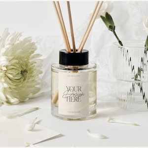 Puede incluir: Un frasco difusor de vidrio transparente con tapa negra y varillas de madera. El frasco tiene una etiqueta color crema con el texto "Your Design Here" en escritura elegante. Flores blancas y un jarrón de vidrio transparente también están en la imagen.