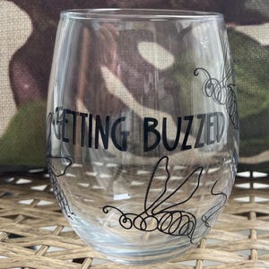 May include: A clear glass wine glass with a black design of bees and the text "Getting Buzzed" printed on the side.