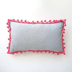 Könnte beinhalten: Ein graues Kissen mit einem rosa gehäkelten Rand. Der Rand hat ein Pompon-Design.