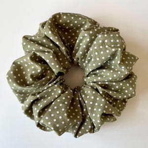 May include: An olive green scrunchie featuring a white polka dot design. This hair accessory is made from soft fabric and is designed to hold hair in a ponytail or bun. A fashionable and practical hair accessory.
