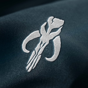 Op de afbeelding: Een wit geborduurd symbool op een donkerblauwe stoffen achtergrond. Het symbool is een gestileerde afbeelding van een Mandalorian helm met een verticale lijn in het midden.