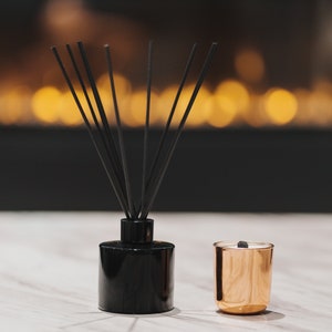 Könnte beinhalten: Ein schwarzer Diffusor für ätherische Öle mit schwarzen Stäbchen und eine kleine goldfarbene Kerze mit einer brennenden Docht. Die Objekte stehen auf einer weißen Marmoroberfläche, mit einem verschwommenen Hintergrund aus warmen, goldenen Lichtern.