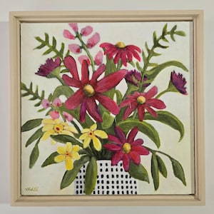 May include: An original floral still life painting. The artwork features a vibrant bouquet of red, pink, and yellow flowers in a white vase with a black grid pattern. The painting is framed in a light wood frame.