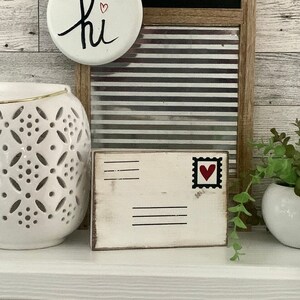 Puede incluir: Linterna de cerámica blanca con borde dorado y diseño recortado. Un letrero de madera blanco con un corazón rojo en un sello y las palabras "hi" en un letrero redondo blanco con un corazón rojo.