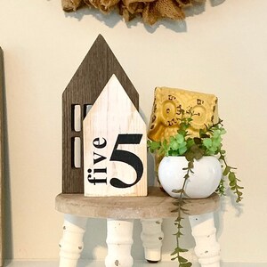 May include: A wooden house-shaped sign with the word "five" and the number "5" painted on it. The sign is sitting on a small wooden stool with white legs. A yellow ceramic owl figurine and a potted plant are also on the stool.