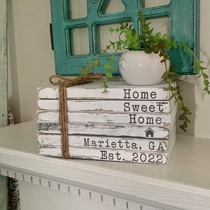 Puede incluir: Una pila decorativa de bloques de madera blancos atados con cuerda, con las palabras "Home Sweet Home" y "Marietta, GA Est. 2022" en letras negras. Una pequeña vasija blanca con vegetación encima, con un marco de ventana turquesa en el fondo.