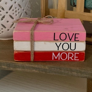 Puede incluir: Tres bloques de madera apilados pintados en rosa, blanco y rojo con las palabras "LOVE YOU MORE" en letras negras y blancas.