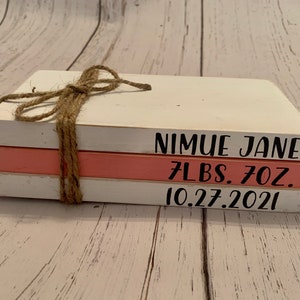 Puede incluir: Una pila de tres libros de madera pintados de blanco con un libro rosa en el medio. El libro superior tiene el nombre "Nimue Jane" escrito en él. El libro del medio tiene "7 lbs. 7 oz." escrito en él. El libro inferior tiene "10.27.2021" escrito en él. Los libros están atados con cuerda.