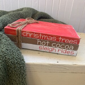 May include: Three stacked wooden blocks with a red, brown, and white paint finish. The blocks are tied together with twine and have the words "christmas trees", "hot cocoa", and "sleigh rides" written on them in white.
